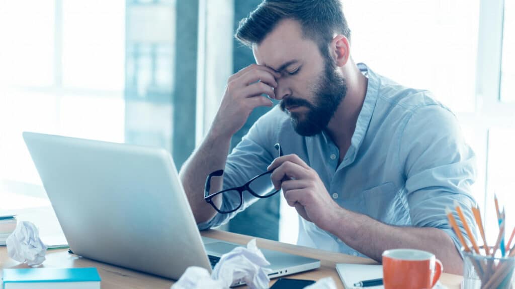Homme stressé assis devant un ordinateur se tenant la tête et ses lunette dans l'autre main.